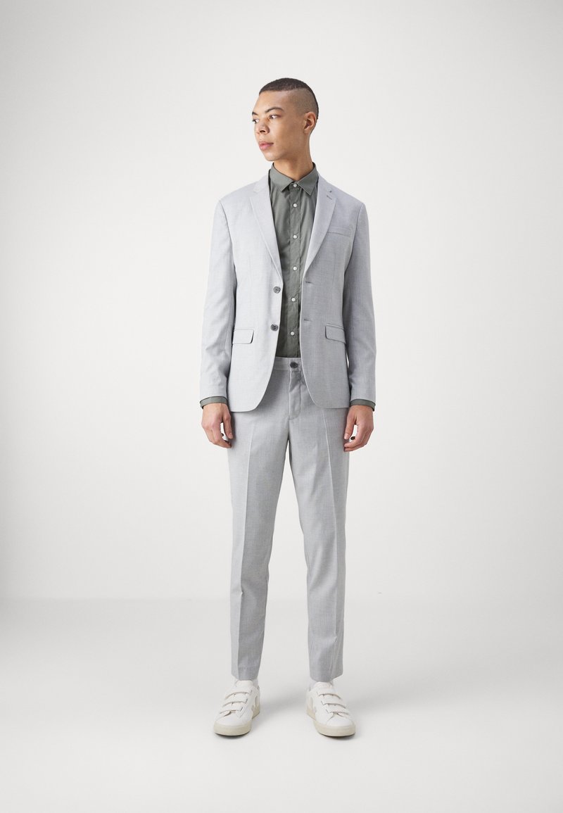 Helles graues Anzug mit einer taillierten Passform, zwei Knöpfen und Klapptaschen, kombiniert mit einem dunkelgrünen Hemd und weißen Sneakern.