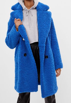 Personne portant un manteau en laine texturé bleu vif par-dessus un sweat à capuche gris et un pantalon noir sur fond blanc.