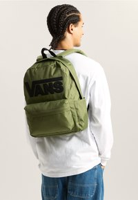 Mochila verde oliva con un gran logo negro de "VANS", que cuenta con correas acolchadas, un bolsillo frontal y un exterior de tela lisa y texturizada.