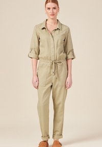 Khaki jumpsuit met een kraag, opgerolde mouwen en een verstelbare tailleband. Beschikt over twee voorzakken en een loose-fit. Zachte stof structuur.