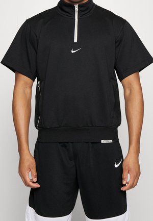 Mężczyzna ubrany w czarny, krótki, półrozpinany pulower Nike oraz czarne krótkie spodenki Nike z białymi logo i akcentami, stojący na jednolitym tle.
