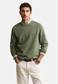 Homme portant un sweat-shirt vert olive sur une chemise rayée, pantalon beige avec ceinture marron et montre-bracelet argentée, mains dans les poches, fond neutre.