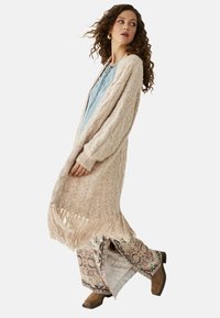 Cárdigan de punto en color beige claro con mangas largas y dobladillo con flecos, combinado con pantalones de chal ligero con estampado y botas marrones hasta el tobillo.