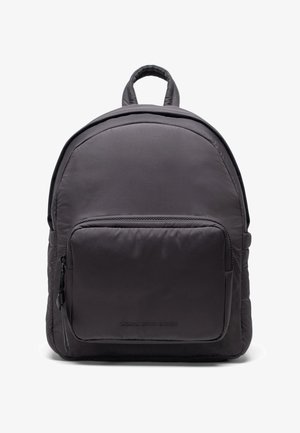 Mochila de nailon gris con forma redondeada, textura acolchada, bolsillo delantero con cremallera y correas ajustables para los hombros. Presenta una sutil marca en la parte inferior.