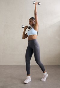 Sostén deportivo azul claro con tirantes ajustables, leggings grises de cintura alta y zapatillas deportivas rosas, sosteniendo mancuernas plateadas por encima de la cabeza.