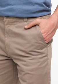 Pantalons en coton beige avec une coupe ajustée, des poches avant et une texture lisse. Couture visible le long des coutures. Main reposant dans la poche.