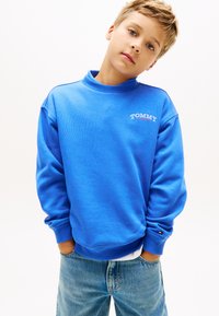 Μπλε φούτερ με υψηλό λαιμό, μακριά μανίκια, ριμπ μανσέτες και μικρό λευκό λογότυπο "TOMMY" στο στήθος. Φορεμένο με ανοιχτόχρωμα μπλε τζιν.