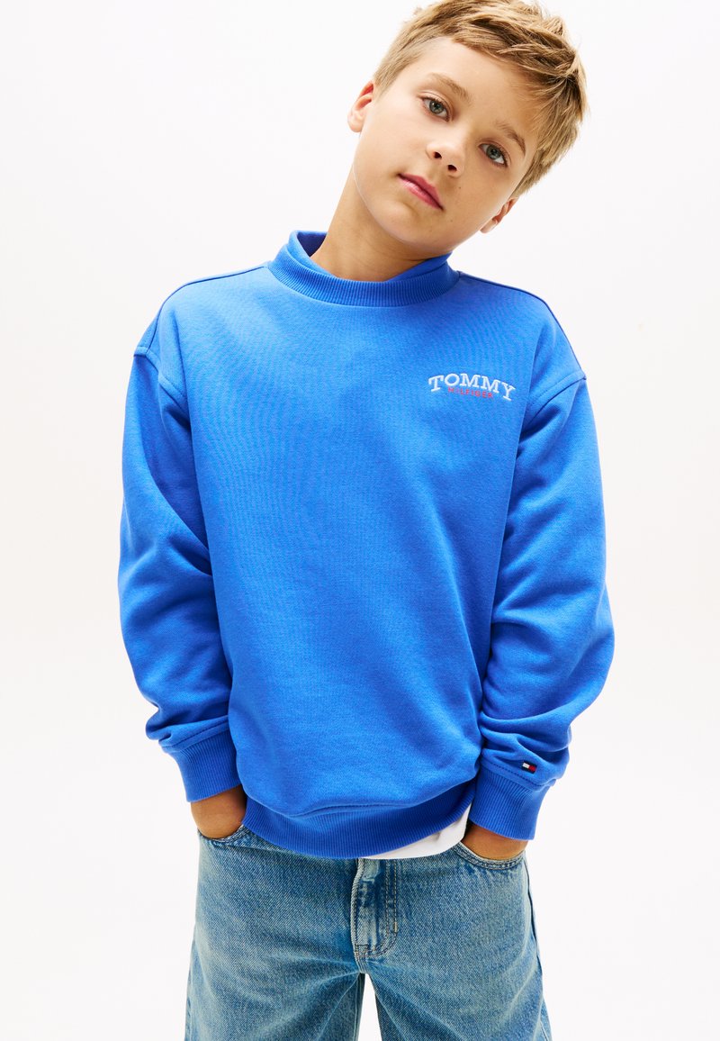 Μπλε φούτερ με υψηλό λαιμό, μακριά μανίκια, ριμπ μανσέτες και μικρό λευκό λογότυπο "TOMMY" στο στήθος. Φορεμένο με ανοιχτόχρωμα μπλε τζιν.