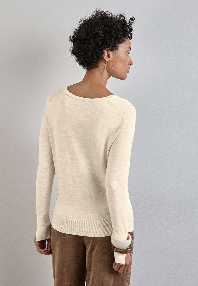 Světle béžový svetr vyrobený z měkkého materiálu, s širokým výstřihem, dlouhými rukávy a pružným lemem, doplněný hnědými kordurovými kalhotami.