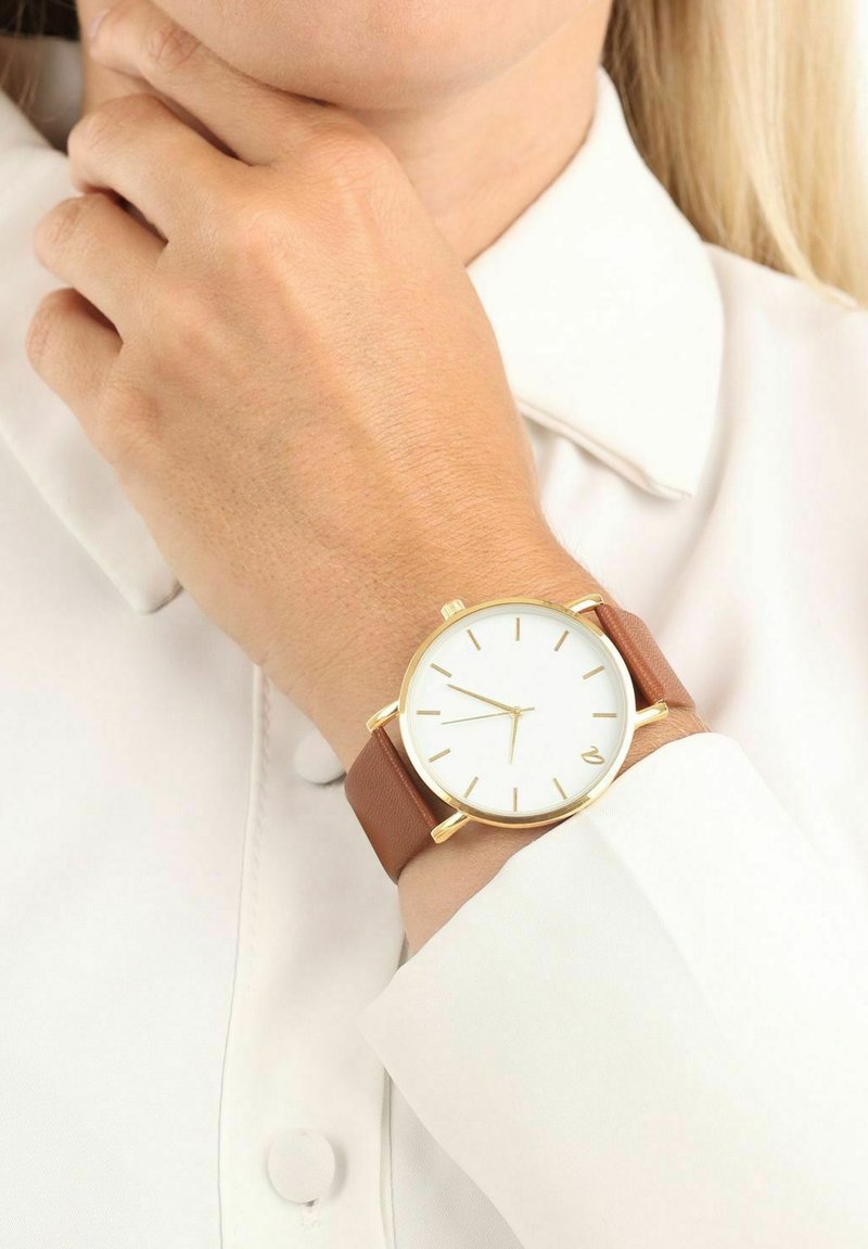 Bruine leren strap horloge met een gouden metalen kast, witte wijzerplaat en gouden uurmarkeringen. Opvallend door het minimalistische ontwerp met een subtiele logo.