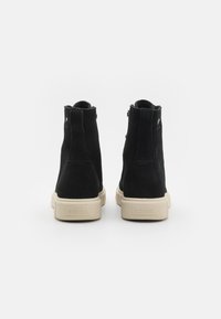 Sorte semska ankelstøvler med beige gummisåler, med rund tå, bakre trekkflik og liten logodetalj nær hælen.