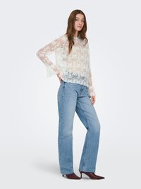 Blouse blanche transparente à motifs floraux avec manches longues, associée à un jean bleu clair à jambes droites et des escarpins bordeaux à bouts pointus.