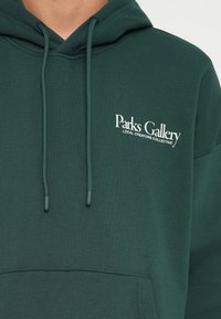 Grüne Kapuzenjacke aus weichem Stoff, mit einer Vorderentasche, verstellbarem Kordelzug und weißem Logotext auf der linken Brust.