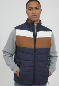 Námornícky puffový vest s horizontálnymi hnedými a bielymi pruhmi, zipsom vpredu, dvoma bočnými vreckami, prešívanou textúrou a kontrastnými sivými rukávmi.