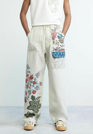 Weite, cremefarbene Hose mit mehrfarbigem Blumenmuster und aufgesetzten Taschen, getragen von einer Person mit den Händen in den Taschen und beigen Turnschuhen.