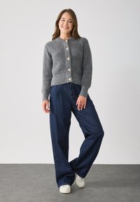 Gray ribbed cardigan with six large cream buttons, paired with loose dark denim pants and white sneakers, set against a light background.