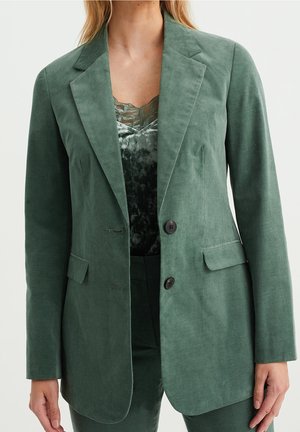 Vrouw draagt een groene fluwelen blazer met twee knopen, bijpassende groene broek en een donker groene fluwelen top met kanten boord eronder.