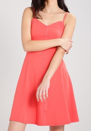 Kobieta w koralowej sukience bez rękawów z guzikami z przodu, pozująca z jedną ręką skrzyżowaną na piersi, a drugą opuszczoną.