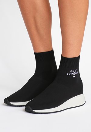 Baskets style chaussette noires avec semelles compensées blanches et inscription « FLY TO LONDON » sur la cheville, portées sur des jambes nues sur fond blanc.