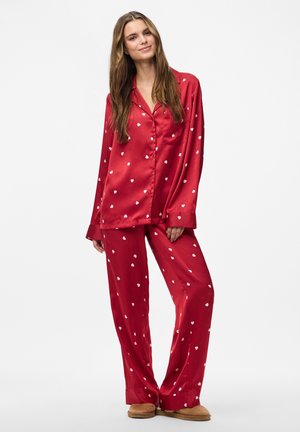 Woman wearing red satin pajama set with white heart prints and tan slippers, standing against a plain white background.