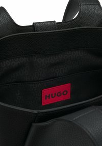 Sort tekstureret lædertaskeindretning med et rødt rektangulært mærke, der viser "HUGO" i sorte bogstaver.