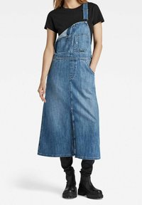 Denim šaty na zavazování s střední délkou, obsahující kapsu na prsou, nastavitelné ramínka a boční rozparek. Nosičná přes černé tričko.