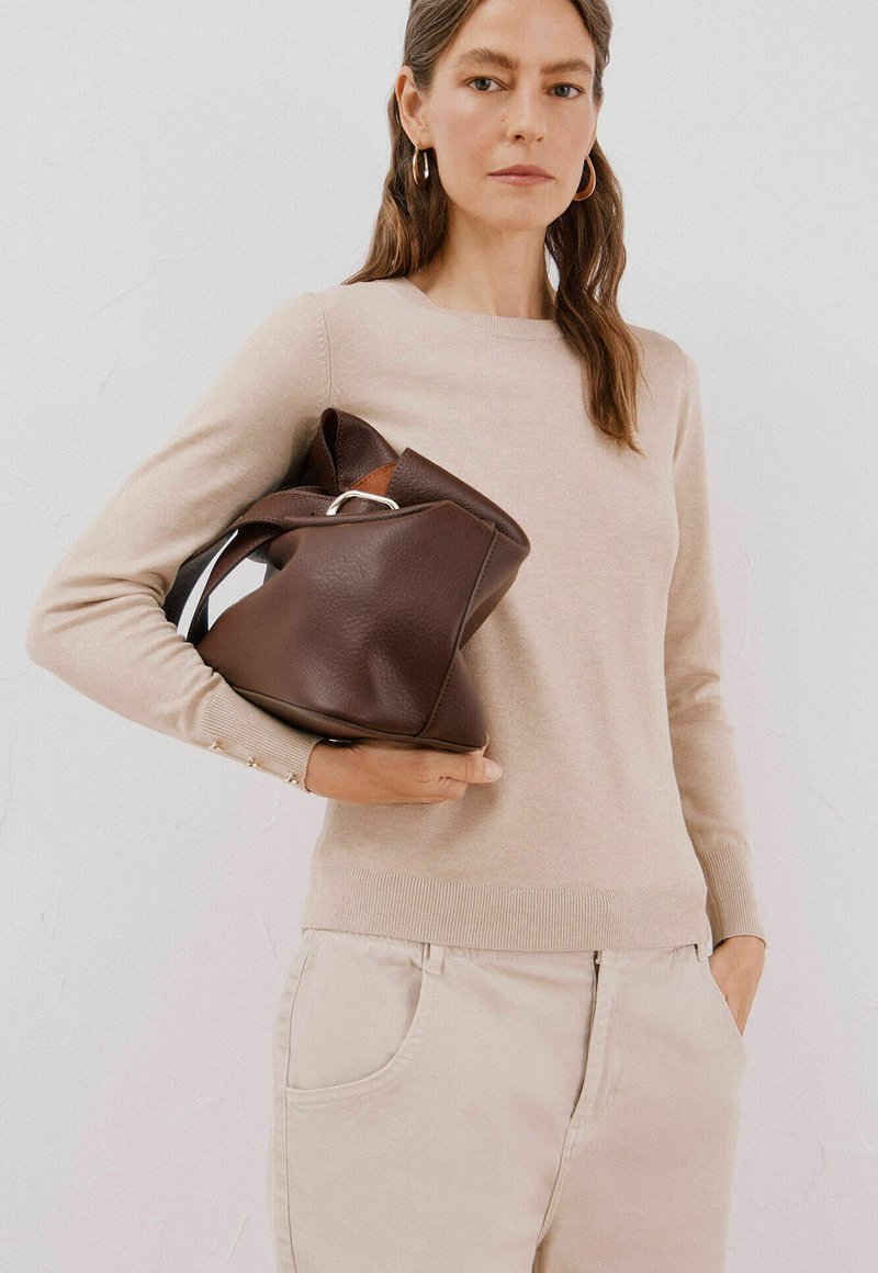 Braune Ledertasche mit strukturiertem Finish, die mit einer Hand gehalten wird. Die Person trägt einen beigen Pullover und helle Hose.