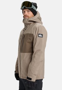 Beige and brown insulated jacket with a zipper closure, adjustable hood, and black accents. Model wearing gloves and ski goggles.