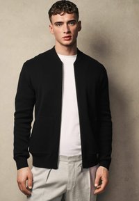 Černý zip-up cardigan s žebrovanou texturou, s dvěma předními kapsami. Nositý přes bílou košili, v kombinaci se světle šedými kalhotami.