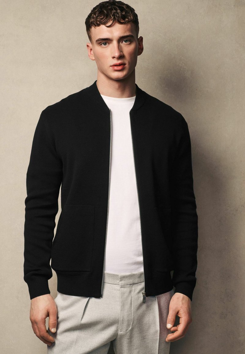 Černý zip-up cardigan s žebrovanou texturou, s dvěma předními kapsami. Nositý přes bílou košili, v kombinaci se světle šedými kalhotami.