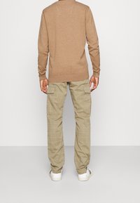 Mężczyzna w beżowym swetrze, khaki w kratkę spodniach cargo z bocznymi kieszeniami i białych sneakersach, stojący na jednolitym tle.