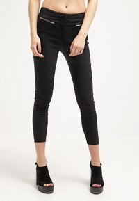 Smala svarta avklippta byxor med diagonala guldfärjade dragkedjor framtill, kombinerade med svarta högklackade öppna ankelstövlar.