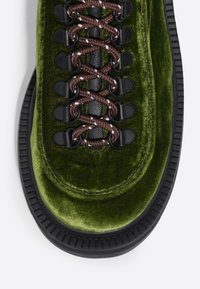 Bota de terciopelo verde con cordones texturizados marrones, ojales negros y una suela de goma negra gruesa con detalles de surcos y costuras.