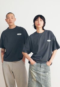 Two individuals wearing oversized dark gray t-shirts. One wears gray sweatpants, the other a snake print skirt. Clean, minimalist style.