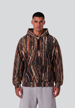 Mann mit kurzen Haaren, der einen braun-schwarz-orangem gemusterten Hoodie und hellgraue Hosen vor einem einfarbigen hellgrauen Hintergrund trägt.