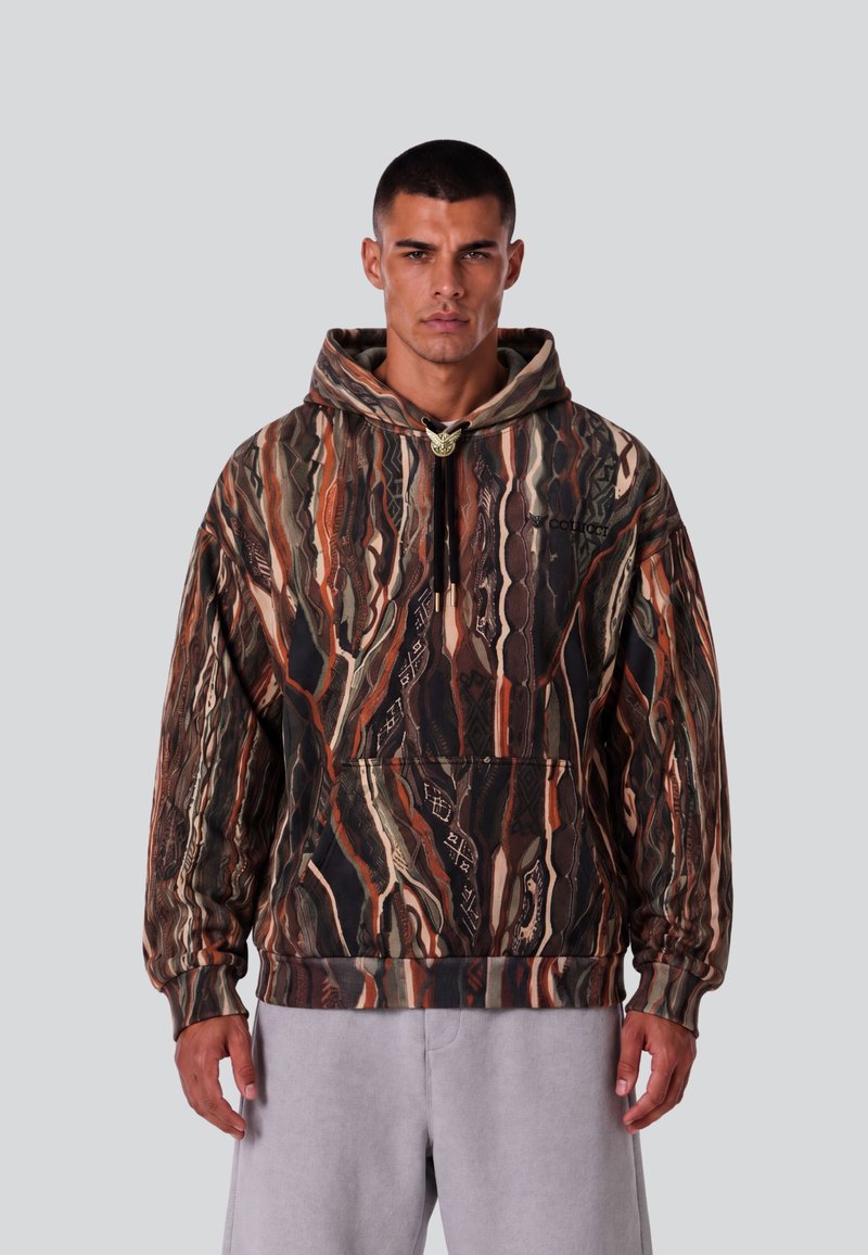 Homme aux cheveux courts portant un hoodie marron, noir et orange à motif, et un pantalon gris clair, sur un fond gris clair uni.