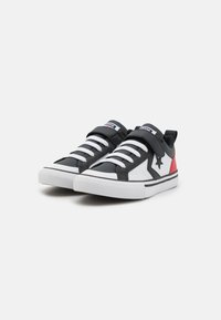 Sneaker in bianco e nero con un accento rosso, dotate di una cintura in velcro e lacci tradizionali, materiali sintetici testurizzati e una suola piatta.