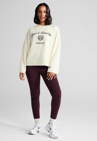 Creme sweater med sort broderet tekst, parret med maroon leggings og hvide sneakers. Sweateren har ribbet kant og en oversized pasform.
