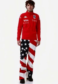 Giacca rossa con zip e loghi; pantaloni con motivo di strisce rosse e stelle bianche su sfondo nero. Tessuto pesante con vestibilità comoda.