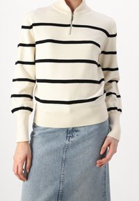 Cremefarbener Pullover mit schwarzen horizontalen Streifen, hohem Kragen und Reißverschluss. Stricktextur mit gerippten Bündchen und Saum. Kombiniert mit einem Jeansrock.