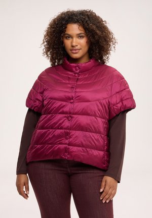 Frau mit lockigem Haar, die eine kurzärmelige, geknöpfte, gesteppte, dunkelrosa Jacke über einem dunkelfarbenen Langarmshirt und dunkler Hose trägt.