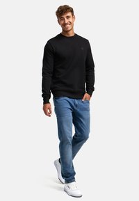 Felpa nera in cotone con scollatura a girocollo, abbinata a jeans denim blu chiaro e sneakers bianche. Il modello sta in piedi con le mani in tasca.
