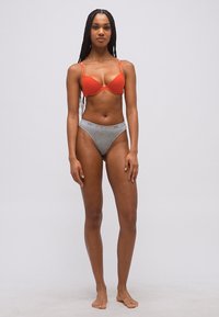 Γυναίκα στέκεται ξυπόλυτη μπροστά σε απλό φόντο, φοράει κόκκινο σουτιέν και γκρι εσώρουχο με τη ζώνη της Emporio Armani, κοιτάζοντας προς τα εμπρός με τα χέρια χαλαρά δίπλα στο σώμα.