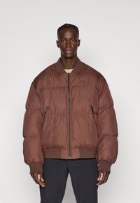 Bruine pufferjack met een ritssluiting aan de voorkant, ribgebreide manchetten en zoom. Bevat een logo patch op de borst en heeft een geborduurde structuur door het geheel.