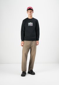 Black sweatshirt with a graphic print, beige trousers, and dark chunky shoes. Red cap features a patterned design. Minimalist background.