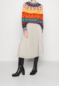 Suéter de lana colorido con patrones en rojo, amarillo, verde y negro, combinado con una falda de pliegues plateada y botas de tacón negras.
