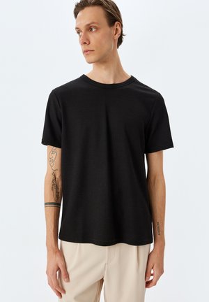 Camiseta negra de manga corta con tejido texturizado, escote redondo y corte relajado, combinada con pantalones de color claro.