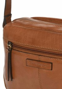 Bruine leren crossbody tas met een ritsluiting, een voorvak met een franjes en een reliëflogo. Soepele textuur, rechthoekige vorm.