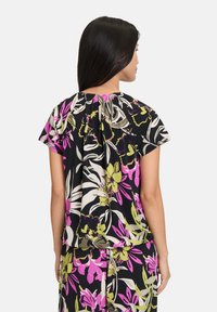 Blouse à manches courtes avec un fond noir, ornée d'un motif floral coloré en rose, vert et blanc. Détail de décolleté froncé à l'arrière.