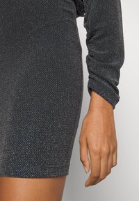 Svart klänning med en glittrig textur, långa ärmar och en figurnära passform. Tyget har glittrande flingor av färg genom hela.